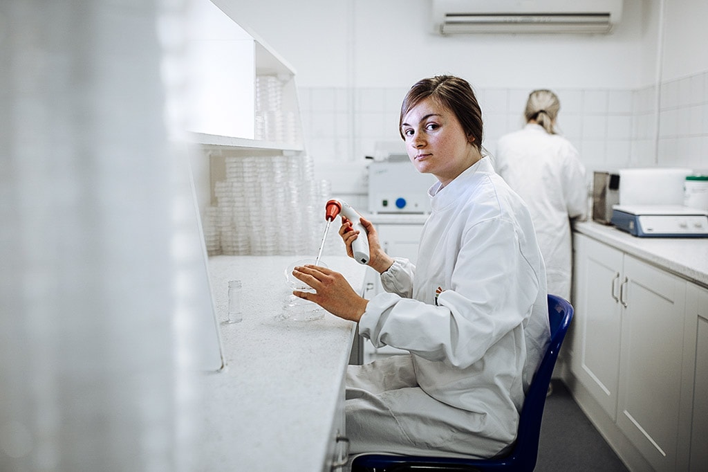 A young female scientist wearing a white lab coat with an embroidered Dawn Farms logo sits with a stack of petri dishes on the counter in front of her.