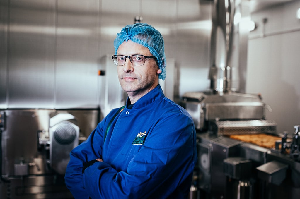 A man wearing Dawn Farms branded blue chef attire and a blue hairnet stands proudly in an industrial kitchen with his arms folded.