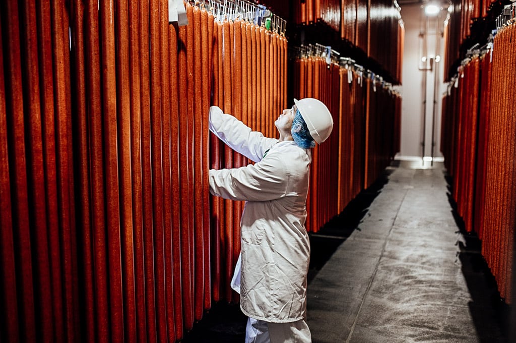 A man in a white coat, hairnet and hard hat examines hanging fermented meats in a large curing room.