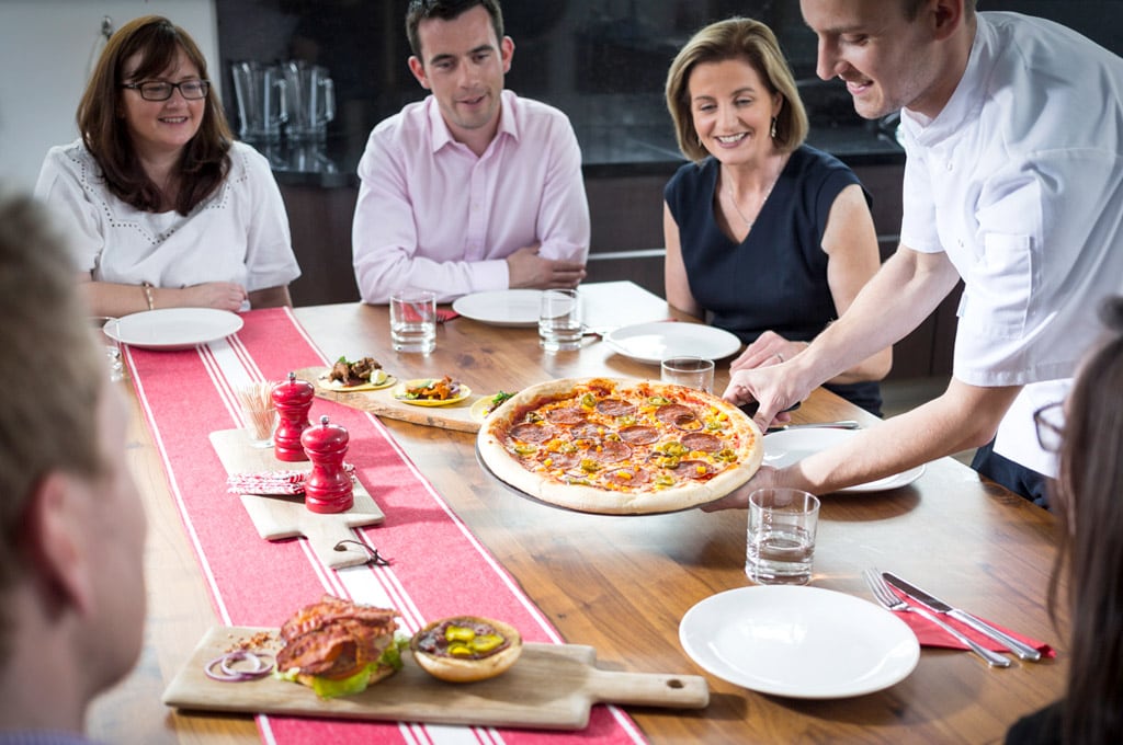 A group of smiling people sit around a table as a chef serves them a pizza topped with Dawn Farms pepperoni.