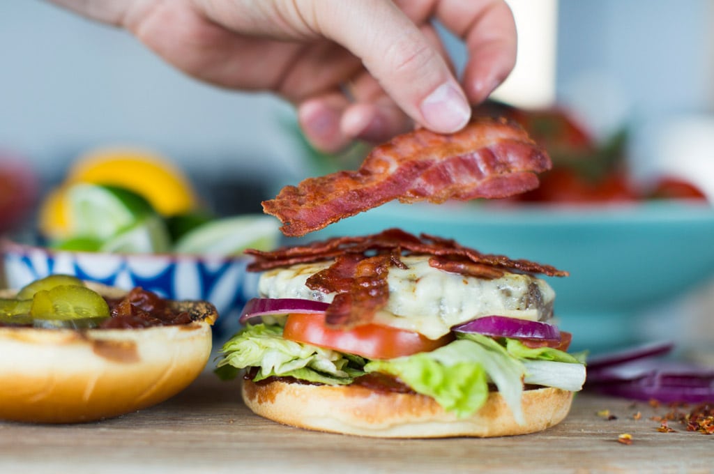 A hand adds a piece of Dawn Farms crispy cooked bacon to a stacked beef burger with cheese, tomato and salad.