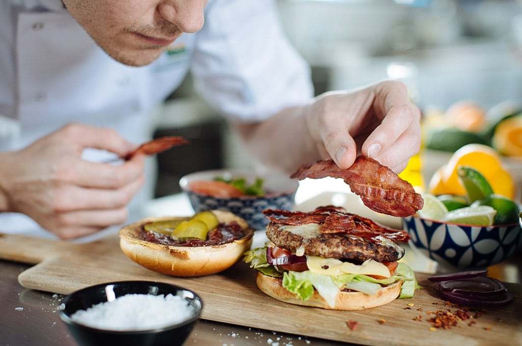 A chef adds a piece of Dawn Farms crispy cooked bacon to a stacked beef burger.