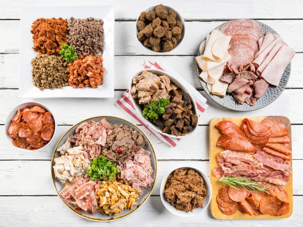 An array of cooked and fermented meat and protein ingredient products – including bacon, ham, chicken, turkey, beef and fermented meats – displayed on a wooden table.