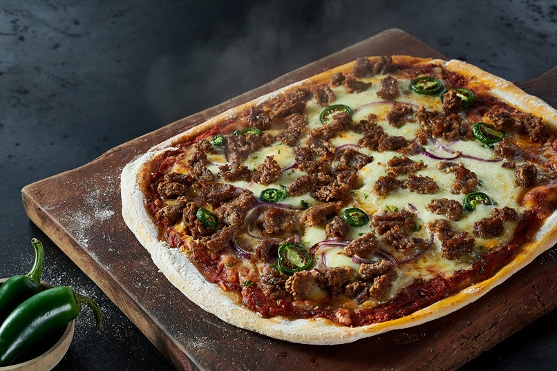 A large, steaming pizza is displayed on a wooden serving board. It is topped with cheese, onions, green tomatoes and Habibi Halal spicy beef chunks.