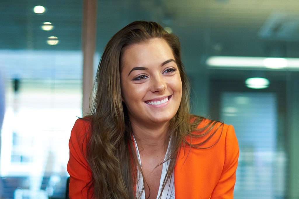 A smiling woman with long hair sits in front of a glass office wall. She wears an orange blazer.