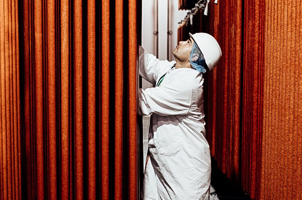 A man in a white coat, hairnet and hard hat examines hanging fermented meats in a large curing room.